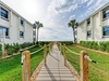 A wooden boardwalk leads between landscaped buildings with palm trees under a partly cloudy sky.