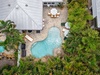 Aerial view showcasing a free-form swimming pool surrounded by lush tropical landscaping, with a paved patio area featuring outdoor dining furniture and lounge chairs near the property entrance.