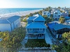 Elevated beach house with blue metal roof sits steps from pristine white sand and turquoise waters in tropical coastal neighborhood.