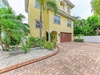 This charming yellow stucco property features Mediterranean-inspired architecture with arched details, surrounded by lush tropical palms and landscaping. A spacious brick-paved driveway welcomes arrivals to the gated entrance.