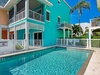 A vibrant turquoise building with white balconies frames the inviting pool area, surrounded by swaying palm trees under a brilliant blue sky, creating a tropical island retreat atmosphere.