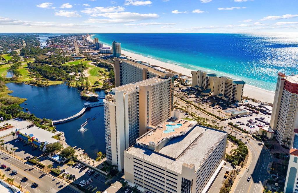 Aerial view of beachfront resort towers along pristine white sand beach with turquoise waters and nearby lake.