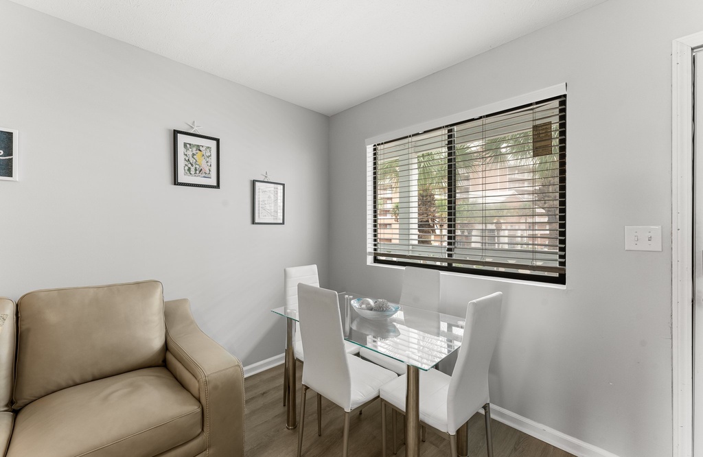 Dining area with a glass table and white chairs next to a window with blinds