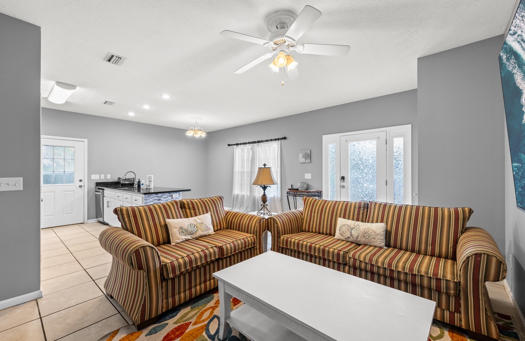 Sink into plush striped sofas in your open-concept living space, where coastal decor, abundant natural light, and a gentle ceiling fan create a breezy, welcoming retreat.
