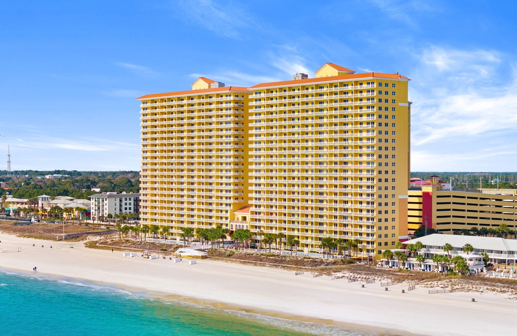 A striking yellow beachfront property stands tall along pristine white sands and turquoise waters, with palm-lined shores and clear blue skies completing this coastal scene.