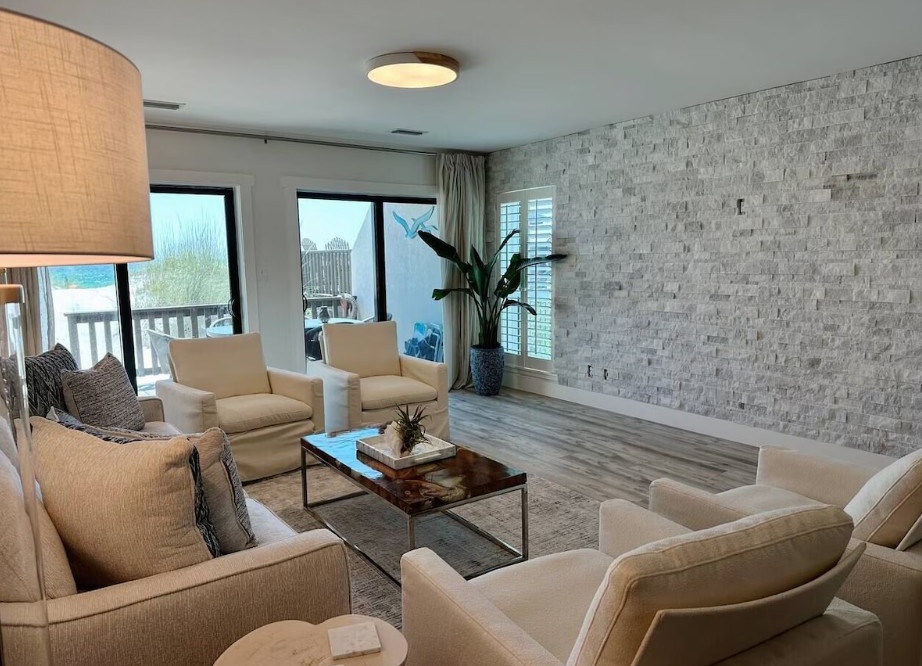 Relax in your airy living room, where coastal light pours through expansive windows, soft neutral furnishings invite conversation, and textured stone walls create a calming, beachy retreat.