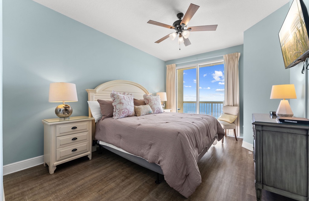Airy bedroom w/ soft blue walls, ceiling fan, & a bed facing the ocean.