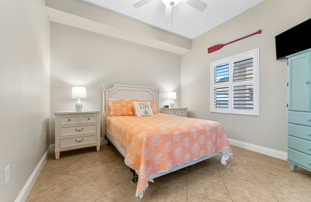 Cozy bedroom with a coral-patterned bedspread and natural light from a window with shutters.