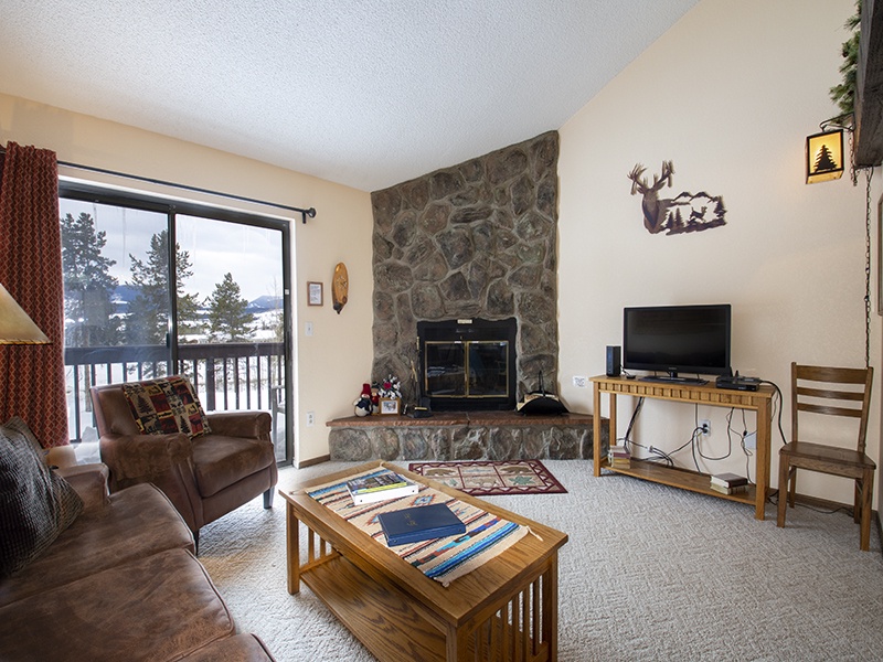 Your cozy mountain retreat awaits with stone fireplace warmth, comfortable seating, and snowy forest views through your private balcony doors.