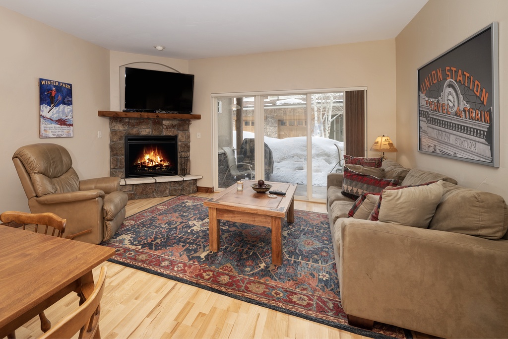 Cozy up in your warm living room with crackling fireplace, comfy seating, and snowy mountain views through sliding doors.