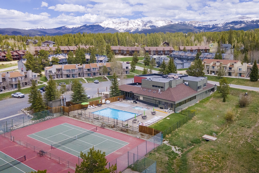 Aerial view of mountain resort community with recreational facilities including tennis court, swimming pool, and modern accommodations set against snow-capped peaks.