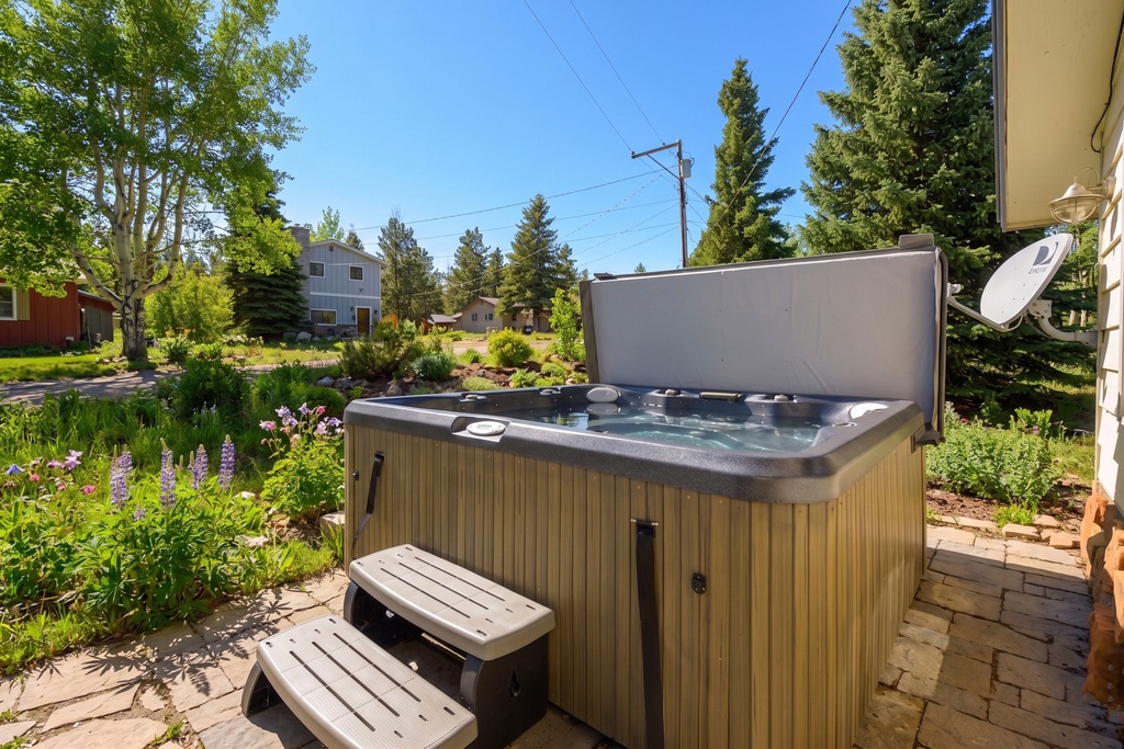 An outdoor jacuzzi set in a tranquil garden setting, framed by lush landscaping, towering evergreens, and vibrant flowers. The property sits in a peaceful residential neighborhood beneath clear blue skies and abundant sunshine.