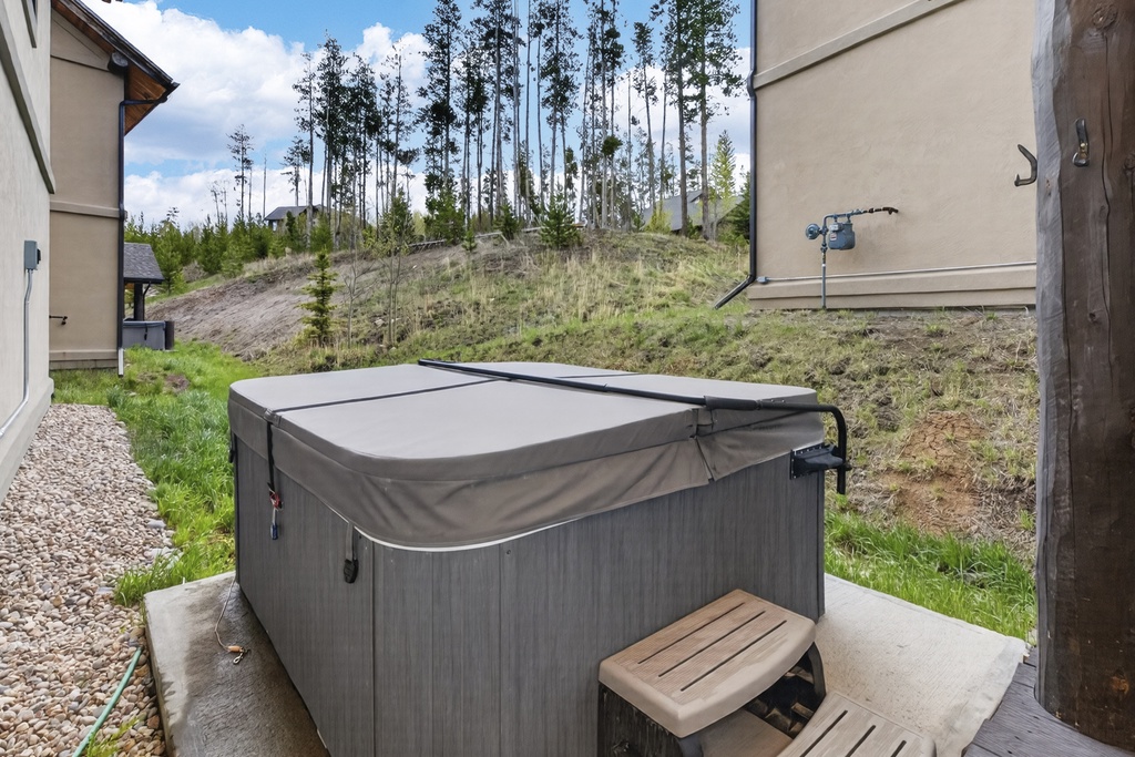 A private hot tub sits on the back patio, offering mountain hillside views. The rental is nestled among tall evergreens in a peaceful natural setting.