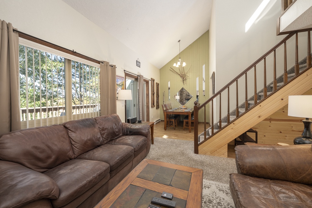 Relax in your spacious open-concept living room featuring plush leather seating, soaring ceilings, and peaceful forest views through large windows.