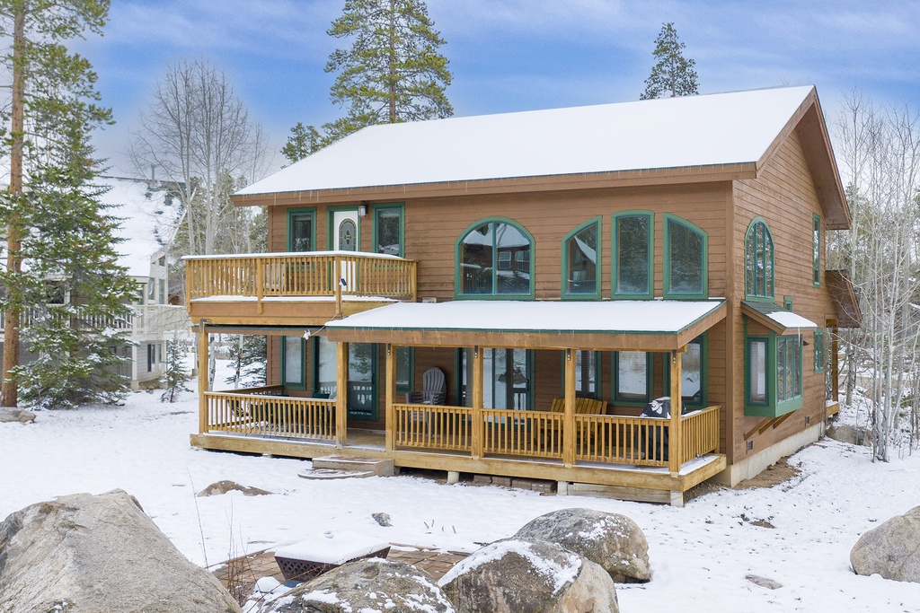 Charming two-story cabin nestled among snow-covered pines, featuring wraparound decks and green-trimmed windows for a perfect winter mountain retreat.