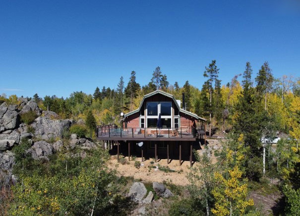 Modern mountain cabin with elevated deck surrounded by pristine summer and towering peaks under clear blue skies.