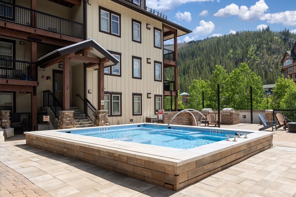 A multi-story property building surrounded by forested mountain views, featuring a raised hot tub and outdoor seating on a paved patio with modern exterior design.