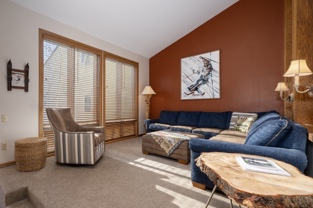 Sink into plush navy sofas where warm sunlight streams through wooden blinds, creating the perfect spot for your mountain retreat relaxation.