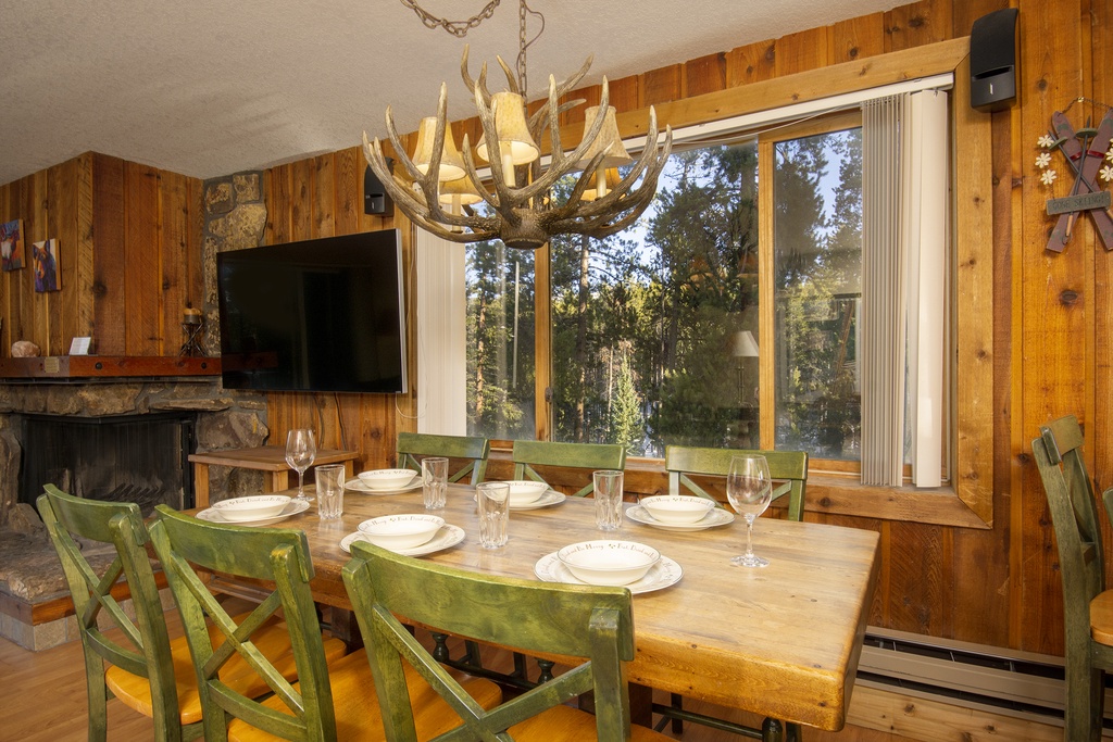Gather around your rustic dining table beneath a stunning antler chandelier, where warm wood surrounds you and forest views create the perfect mountain retreat atmosphere.