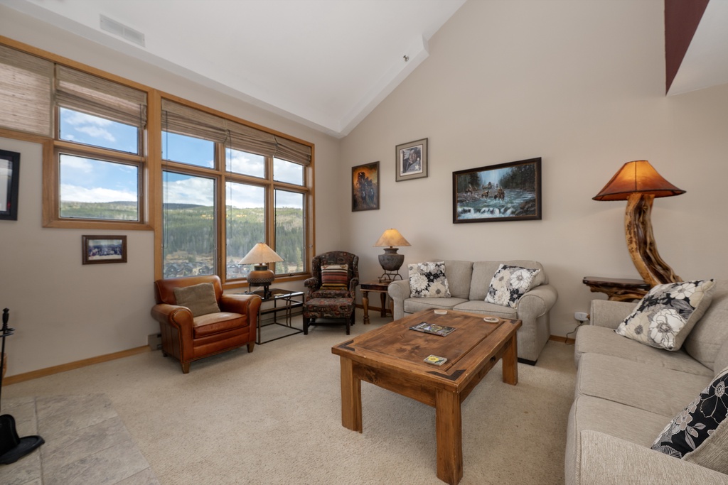 Relax in your mountain retreat's bright living space, where floor-to-ceiling windows frame stunning forest views and cozy furnishings invite you to unwind.