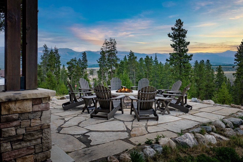 Elevated stone patio encircles a central fire pit surrounded by Adirondack chairs, offering sweeping mountain and valley vistas framed by evergreen forest.