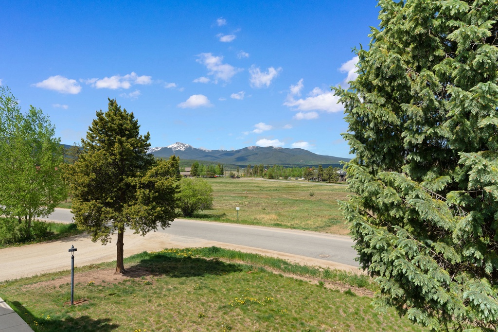 A scenic mountain landscape with lush greenery, open fields, and quiet residential streets framing distant peaks under a clear blue sky.