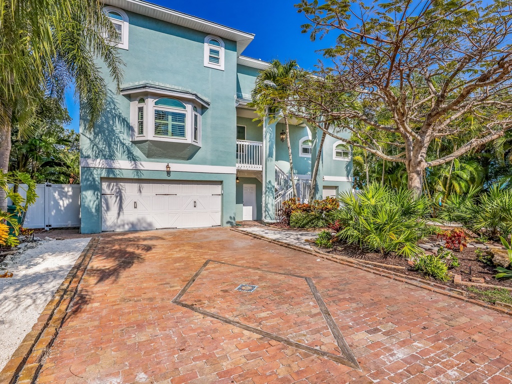 Stunning turquoise coastal home surrounded by lush tropical landscaping and elegant brick driveway under brilliant blue skies.