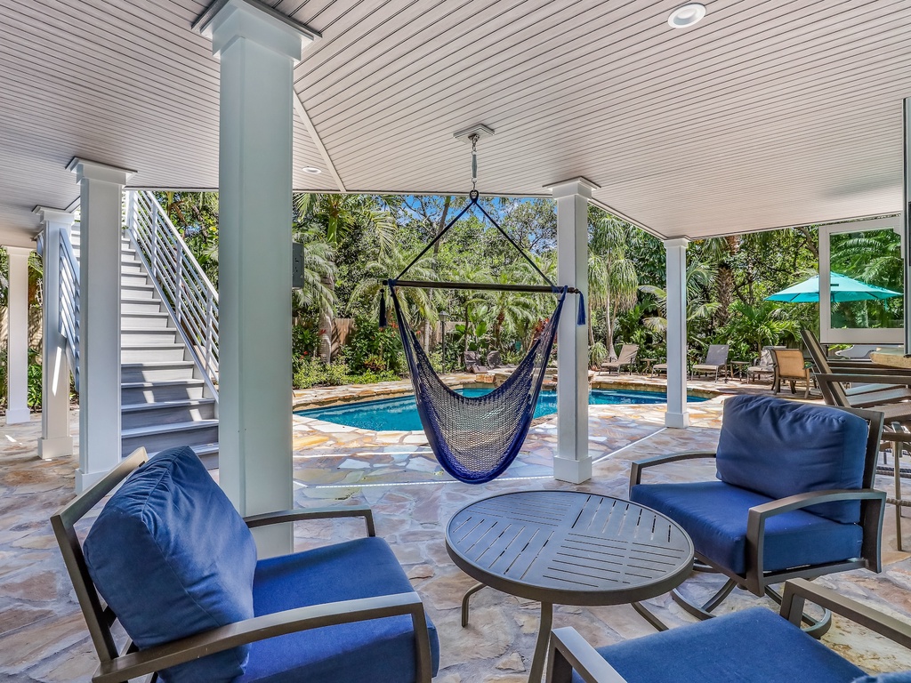 Unwind in your covered patio with plush blue seating, a swing chair, and pool views surrounded by tropical greenery.