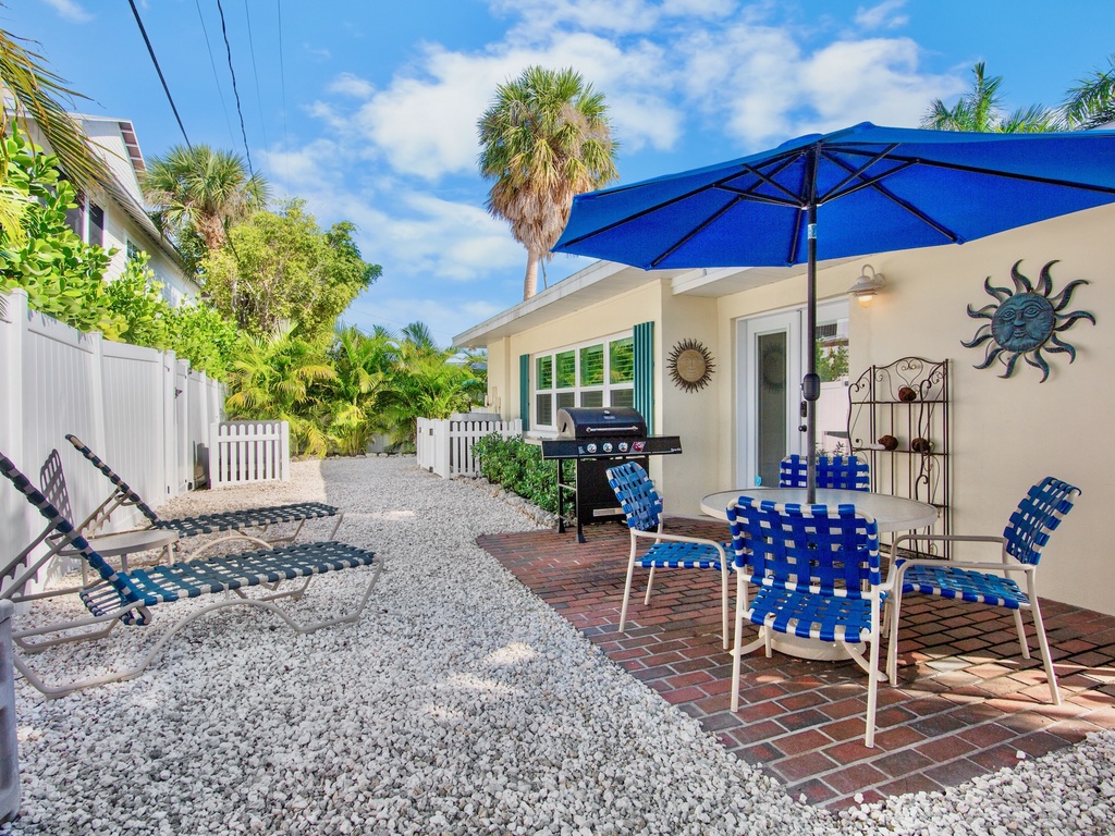 Your private outdoor oasis features a BBQ grill, comfortable seating under a bright blue umbrella, and lounge chairs on tropical landscaping - perfect for relaxing and dining outdoors.