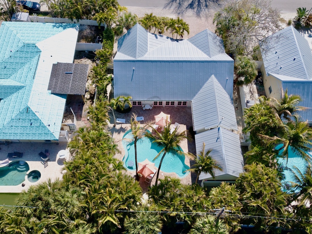 Aerial view of tropical resort buildings with distinctive white roofs surrounded by palm trees and multiple swimming pools.