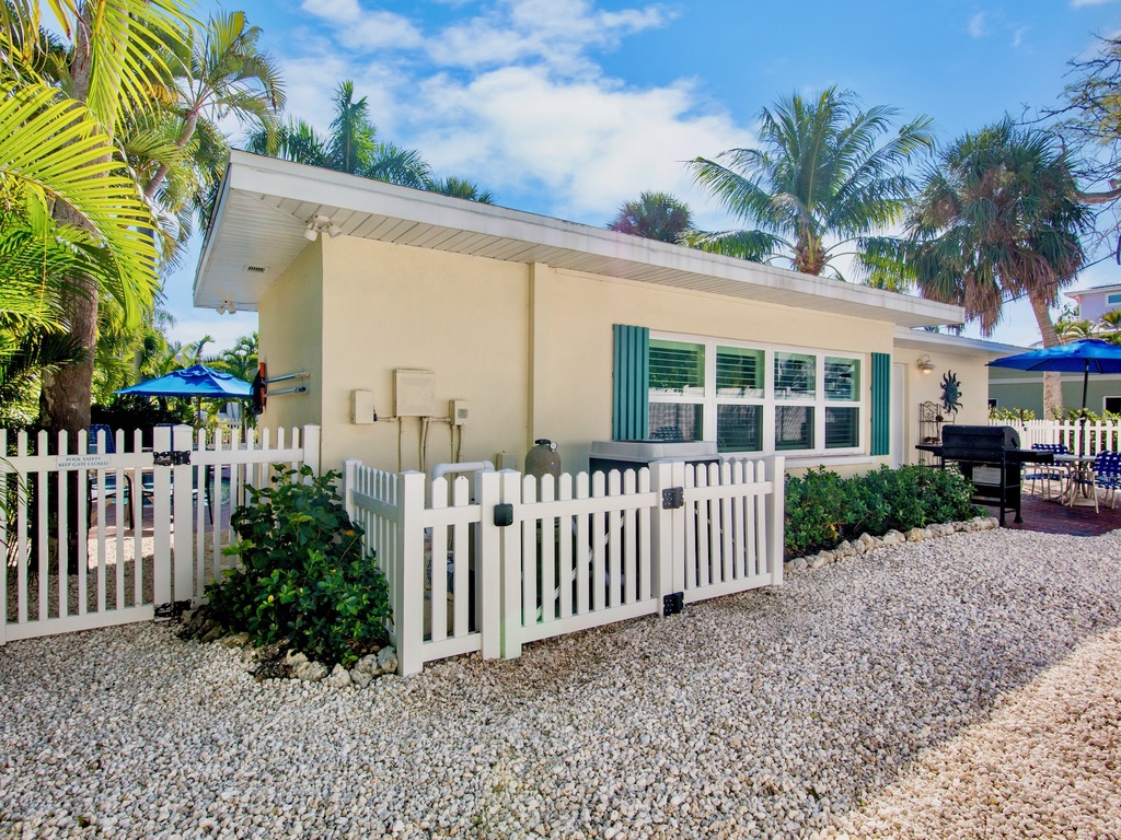Charming beachside cottage with white picket fence surrounded by tropical palm trees and landscaped gardens in a sunny coastal setting.