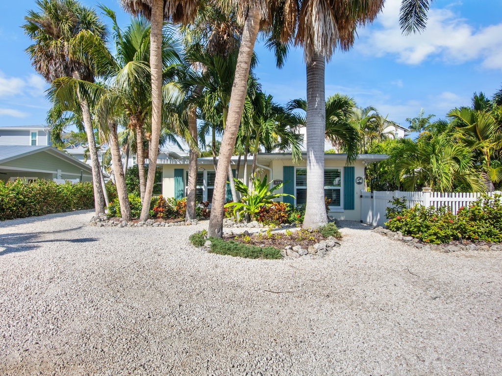 Tropical property entrance surrounded by swaying palm trees and lush landscaping with convenient gravel parking area.