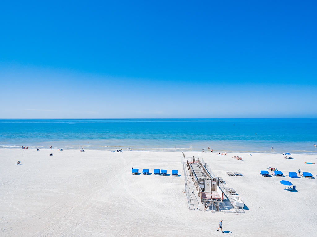 Pristine white sand beach stretches along crystal-clear turquoise waters under brilliant blue skies, with beach chairs and boardwalk access for guests.