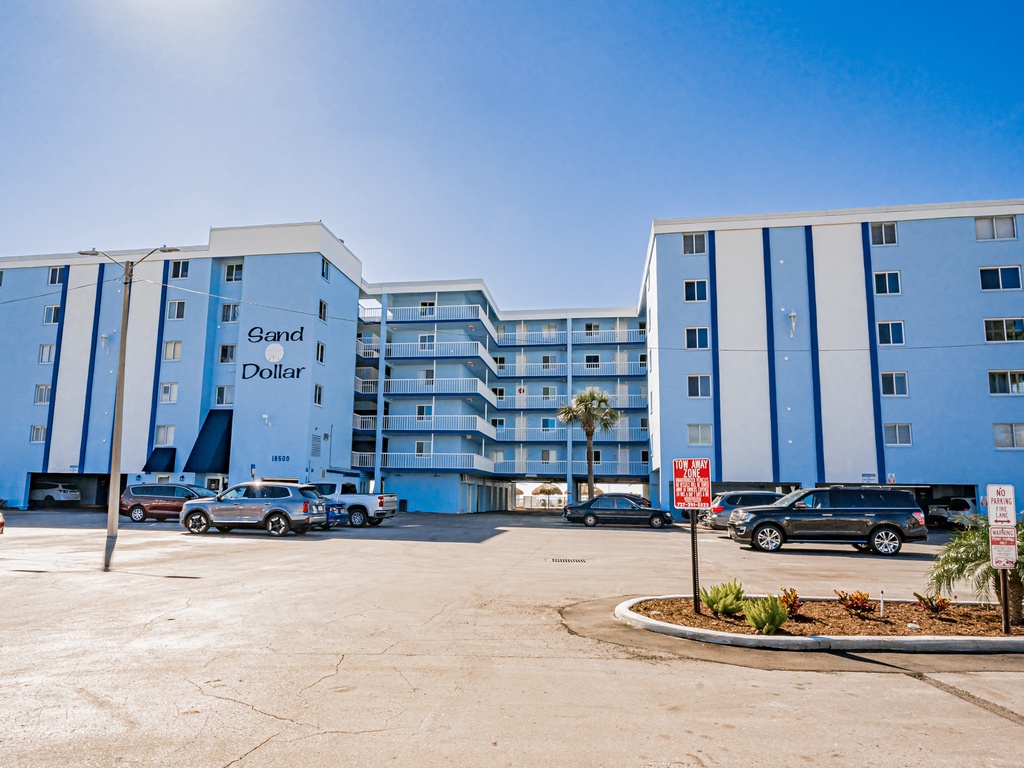 Sand Dollar condominium complex features multiple blue buildings with convenient parking and tropical landscaping under clear blue skies.