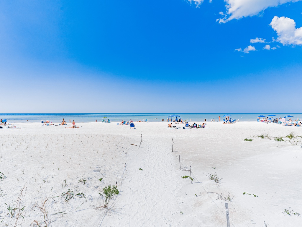 Pristine white sand beach stretches endlessly under brilliant blue skies, with gentle waves and scattered beachgoers enjoying the perfect coastal setting.