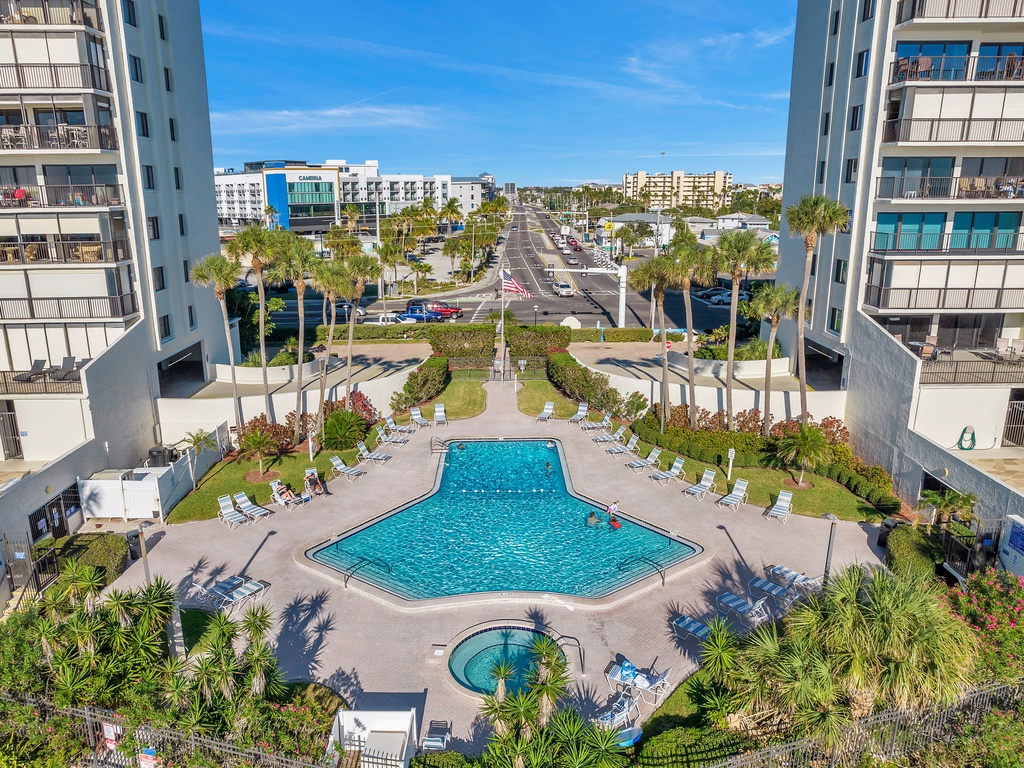 Aerial view of a modern resort complex with swimming pool and landscaped gardens, surrounded by mid-rise buildings and palm trees.