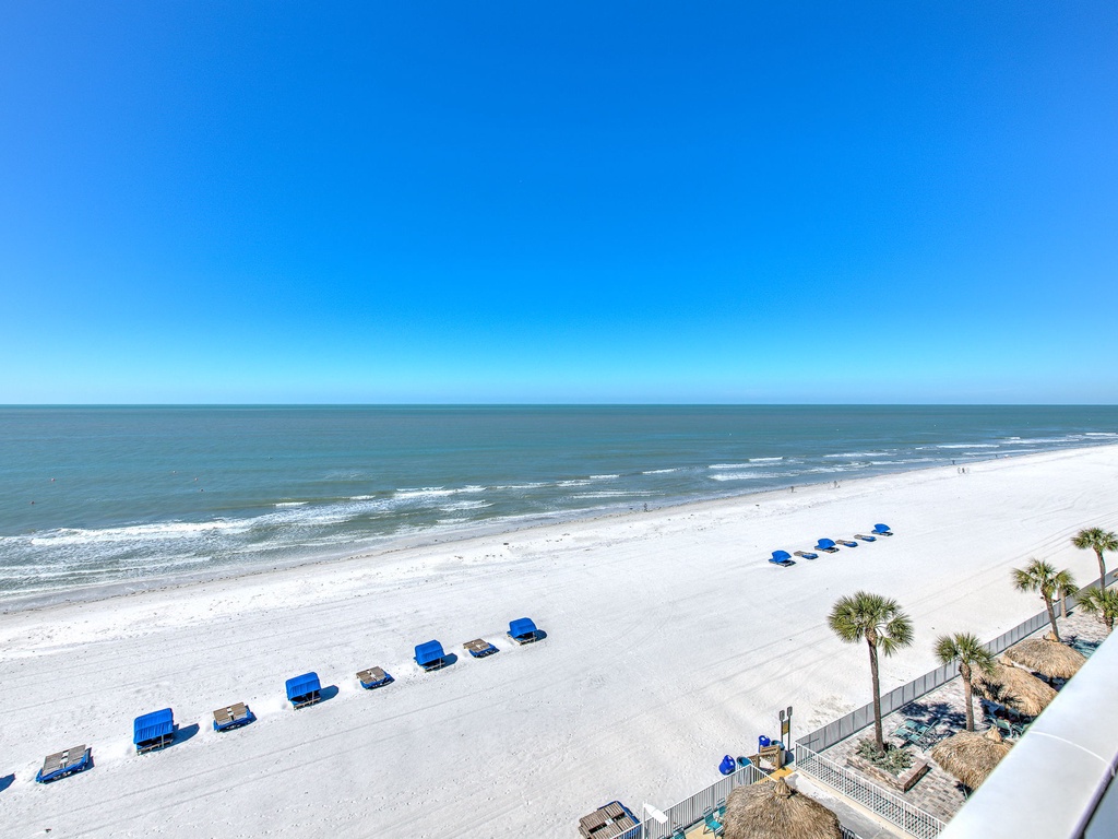 Pristine white sand beach stretches along crystal-clear waters under brilliant blue skies with rental umbrellas and palm trees dotting the shoreline.