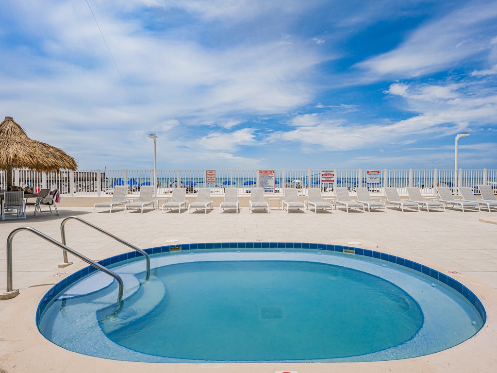 Beachfront pool area featuring a tiki hut, lounge chairs, and clear blue waters with direct beach access beyond the fence.
