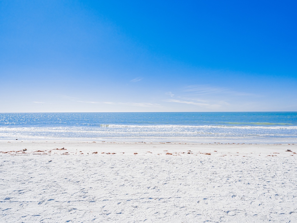 Pristine white sand beach stretches endlessly under brilliant blue skies, with gentle waves rolling onto the peaceful shoreline.
