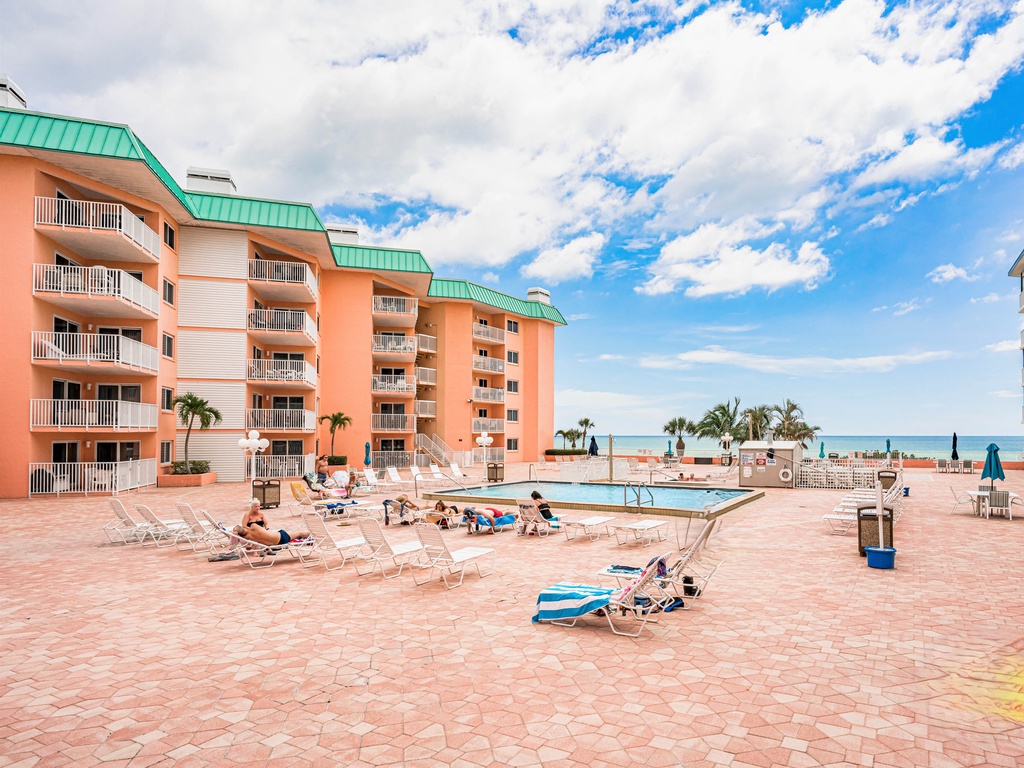 Beachfront resort property with coral-colored buildings, green roofs, pool area, and direct beach access under sunny skies.