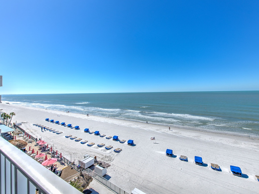 Pristine white sand beach stretches endlessly beneath brilliant blue skies, with organized beach chair rentals and beachfront property visible in the scene.