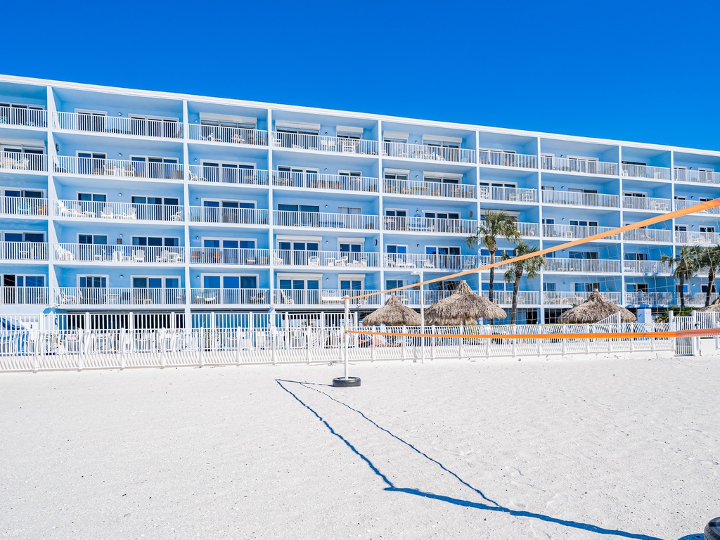 Beachfront property with white sand, volleyball court, and thatched umbrellas offering direct beach access under brilliant blue skies.