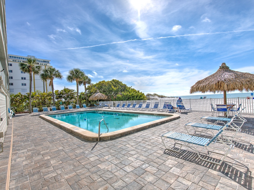 Beachfront pool area with tiki umbrella, lounge chairs, and palm trees under bright blue skies near the white sand beach.