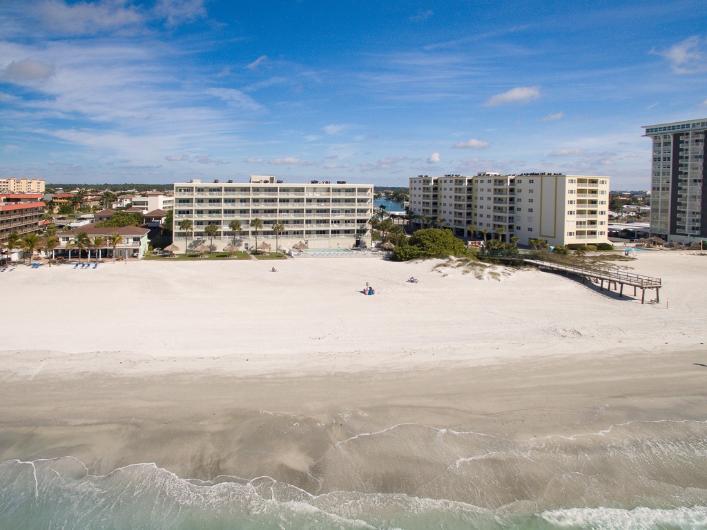 Aerial view of beachfront property buildings along pristine sandy shoreline with clear blue sky and gentle waves.