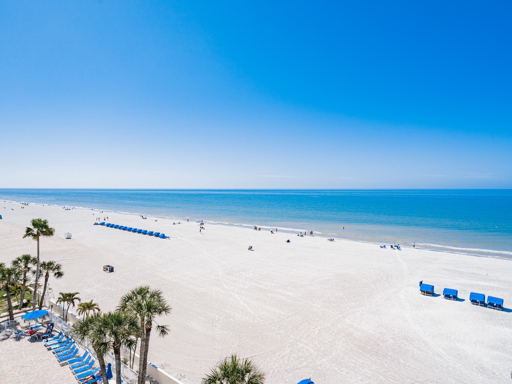 Pristine white sand beach stretches along turquoise waters under clear blue skies, with palm trees and beach chairs dotting the shoreline.
