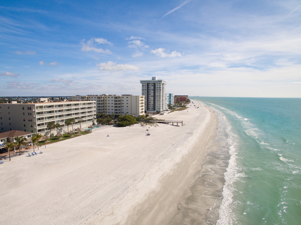 Aerial view showcases pristine white sand beach stretching along turquoise waters, with oceanfront hotels and condominiums lining the shore.