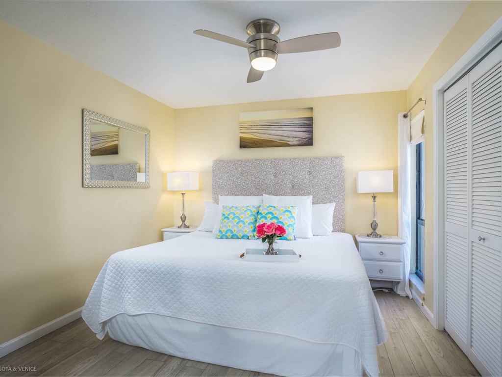 Drift off under warm yellow walls and gentle ceiling fan breezes in your serene bedroom sanctuary with ocean artwork creating peaceful vibes.