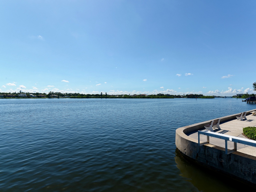 Waterfront seating area overlooking a peaceful lake with distant residential shoreline and blue sky.