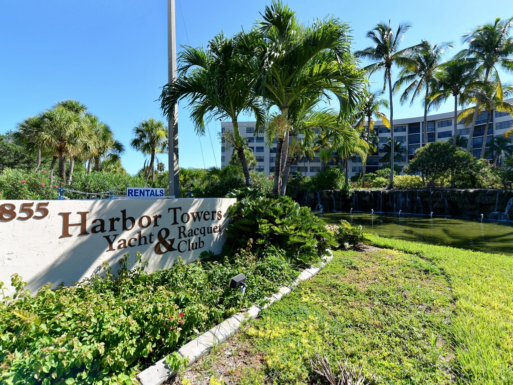 Harbor Towers Yacht & Racquet Club entrance surrounded by lush tropical landscaping and towering palm trees under brilliant blue skies.