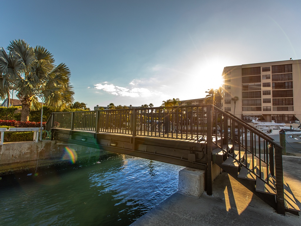 Waterfront bridge connects you to the marina area with tropical palms and nearby property buildings in warm sunlight.